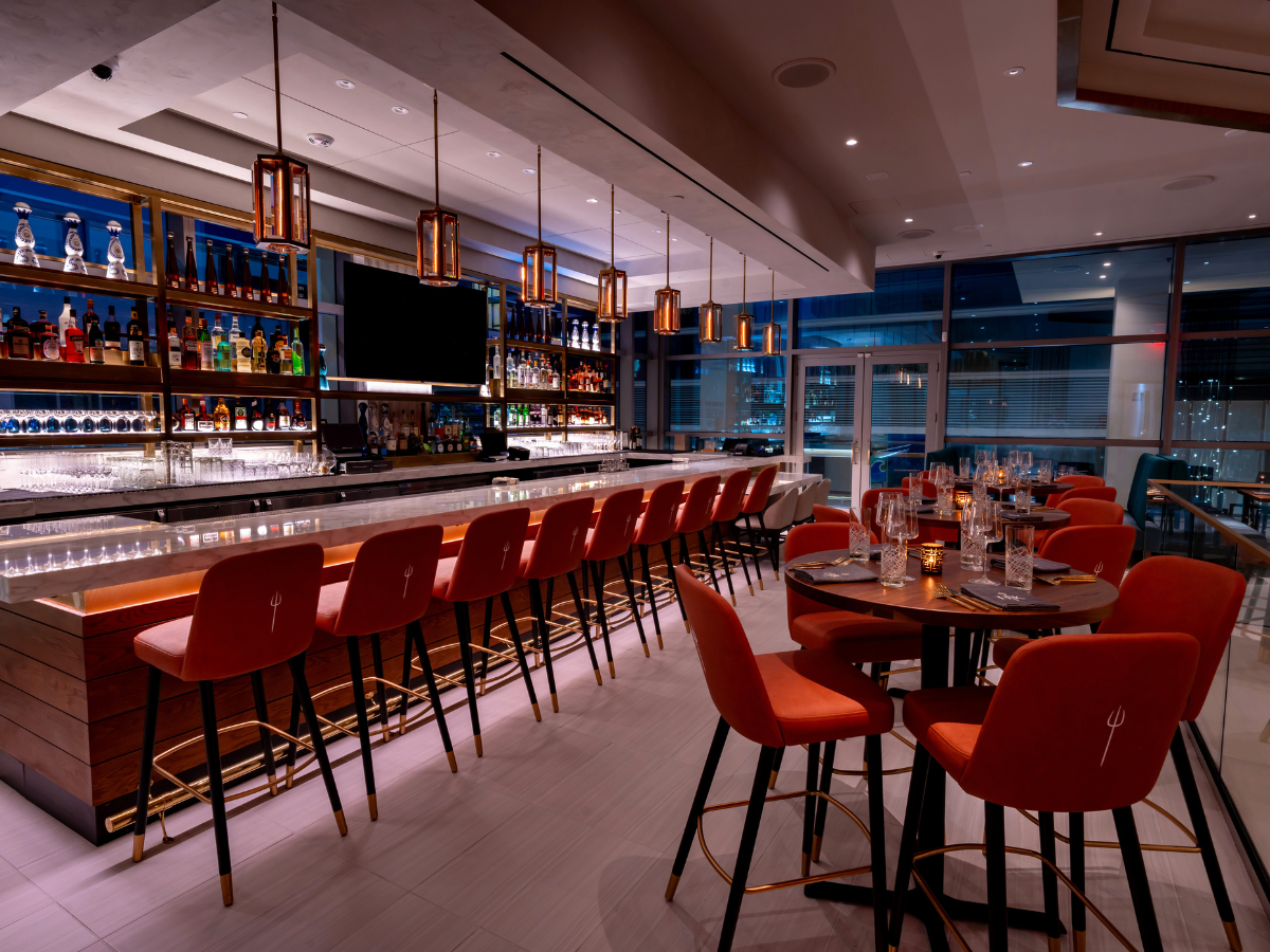 A modern bar interior with orange stools, a long marble-topped counter, and tables set for dining.