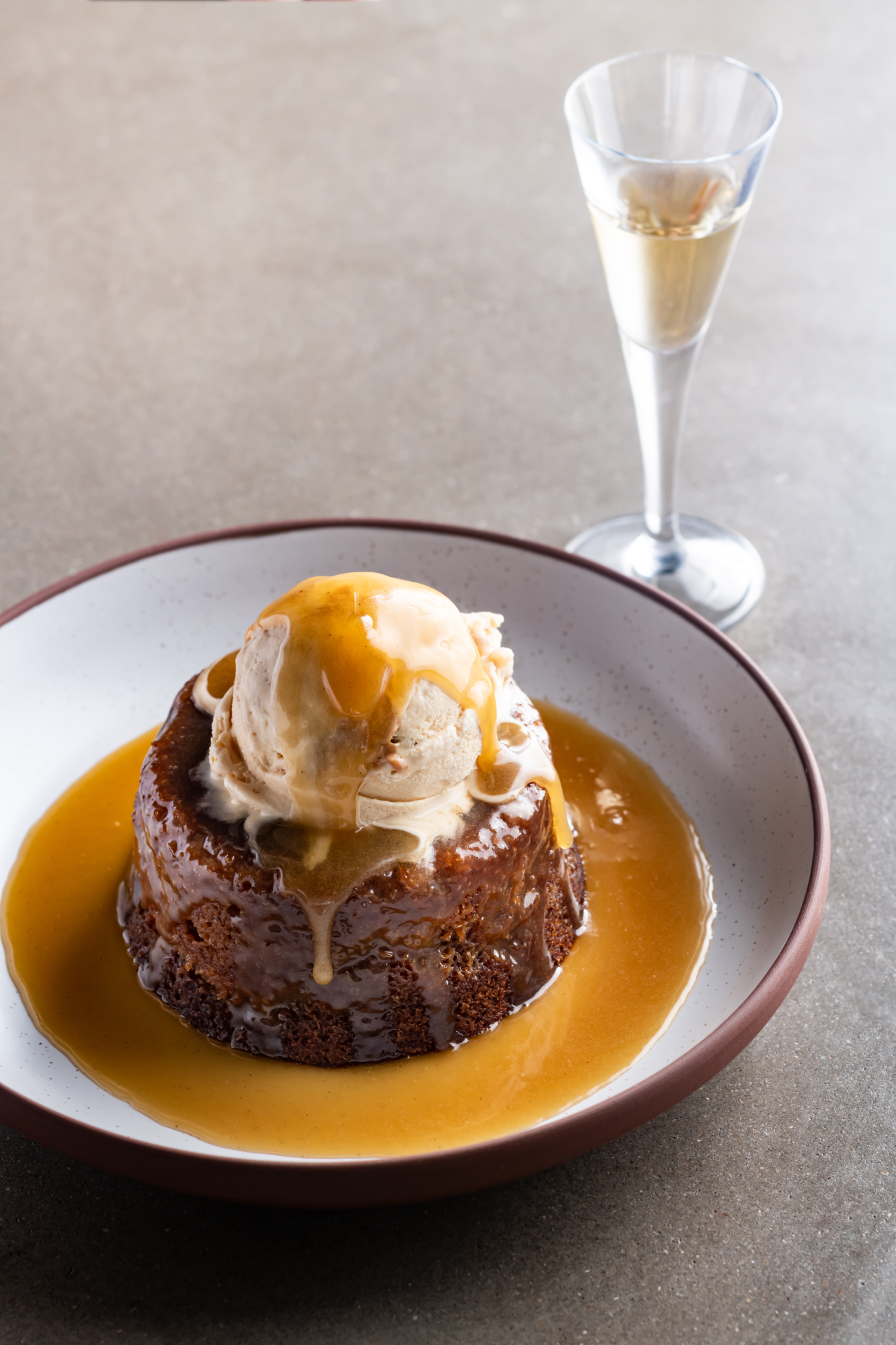 Sticky toffee pudding topped with a scoop of ice cream and caramel sauce, served with a glass of dessert wine.