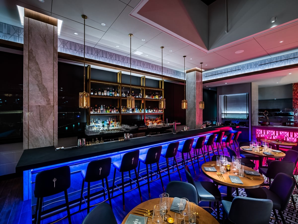 Modern restaurant bar with blue neon under-lighting, a well-stocked spirits shelf, and circular dining tables.