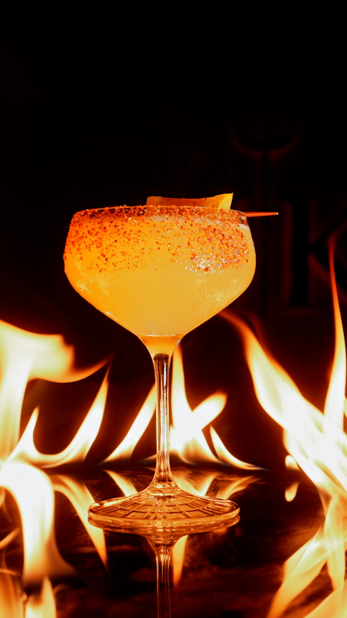 A cocktail in a stemmed glass with a chilli-salted rim, surrounded by flames.