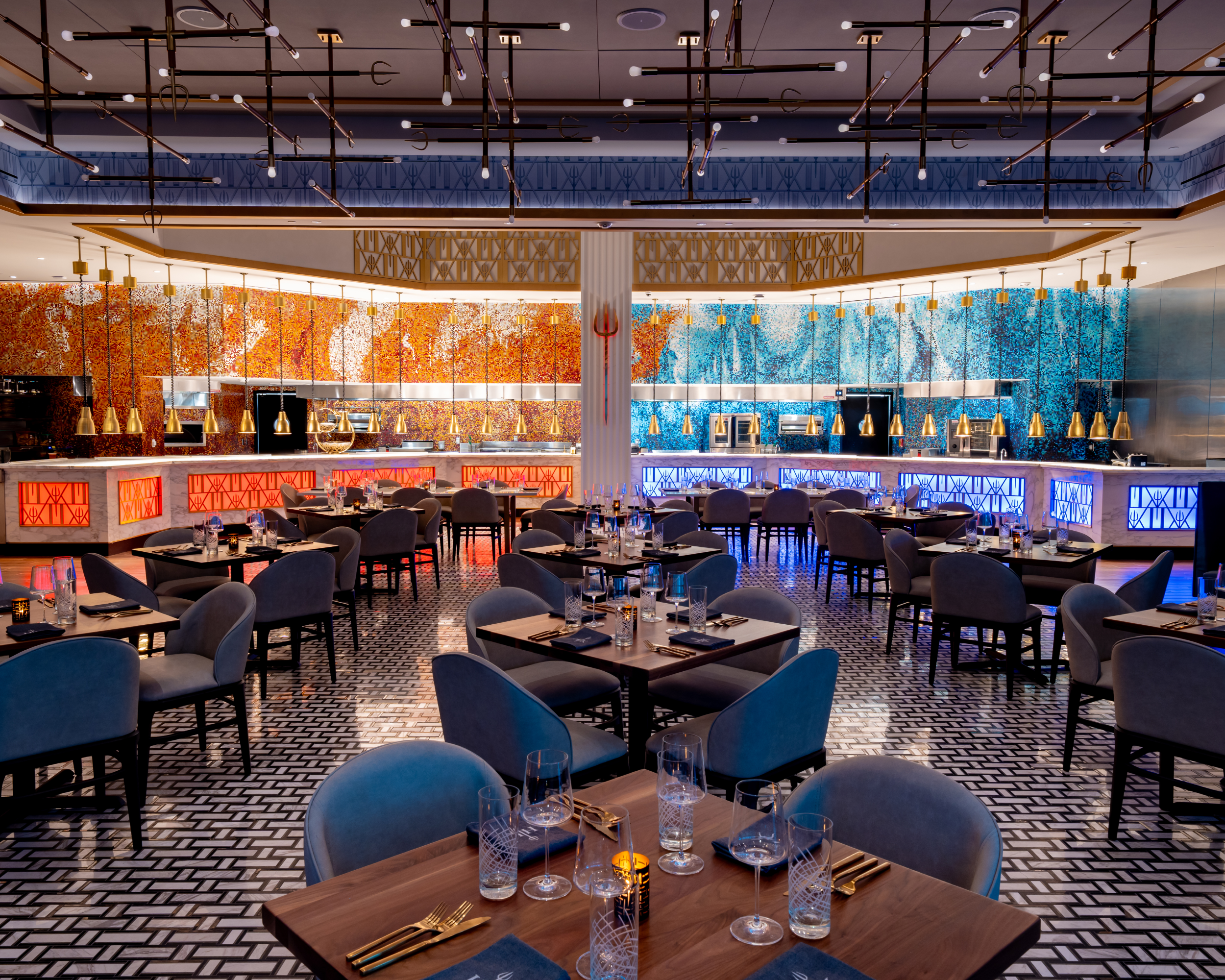 A dining room with many tables and chairs, featuring a long bar backed by colorful orange and blue mosaic tile walls.
