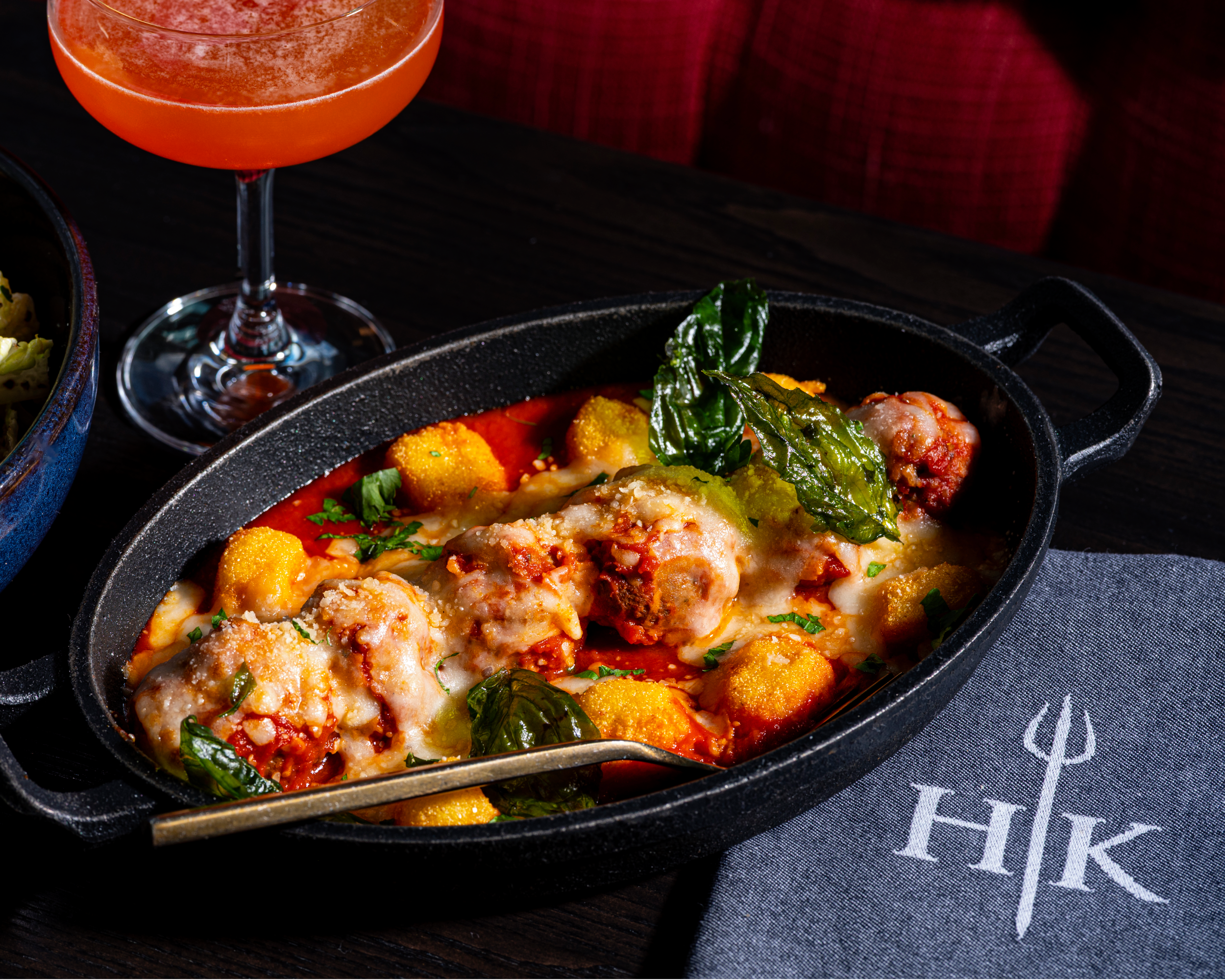 A cast-iron frying pan of meatballs and polenta in tomato sauce, served next to a cocktail and a Hell’s Kitchen serviette.