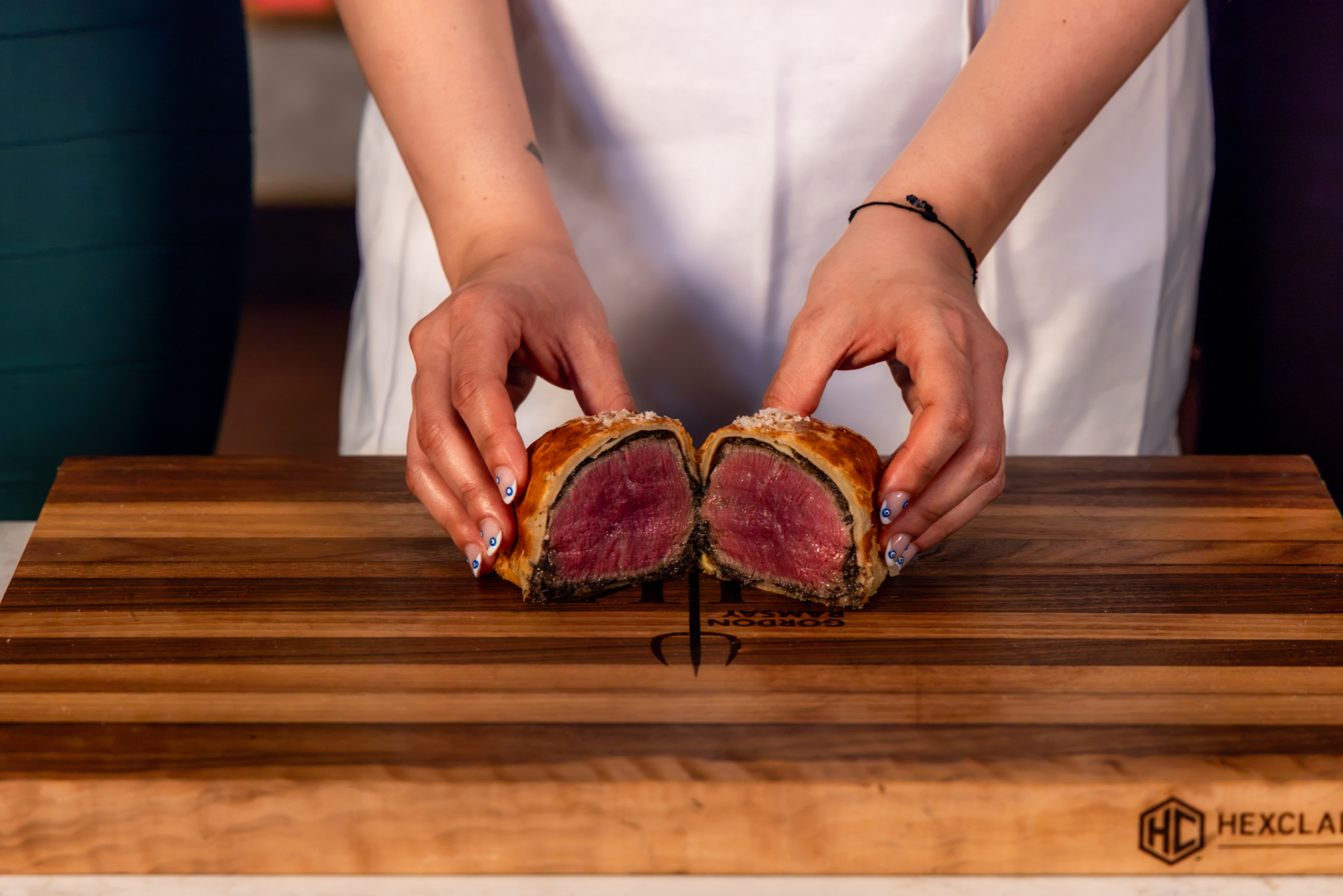 Hands pulling apart a halved Beef Wellington on a wooden chopping board.