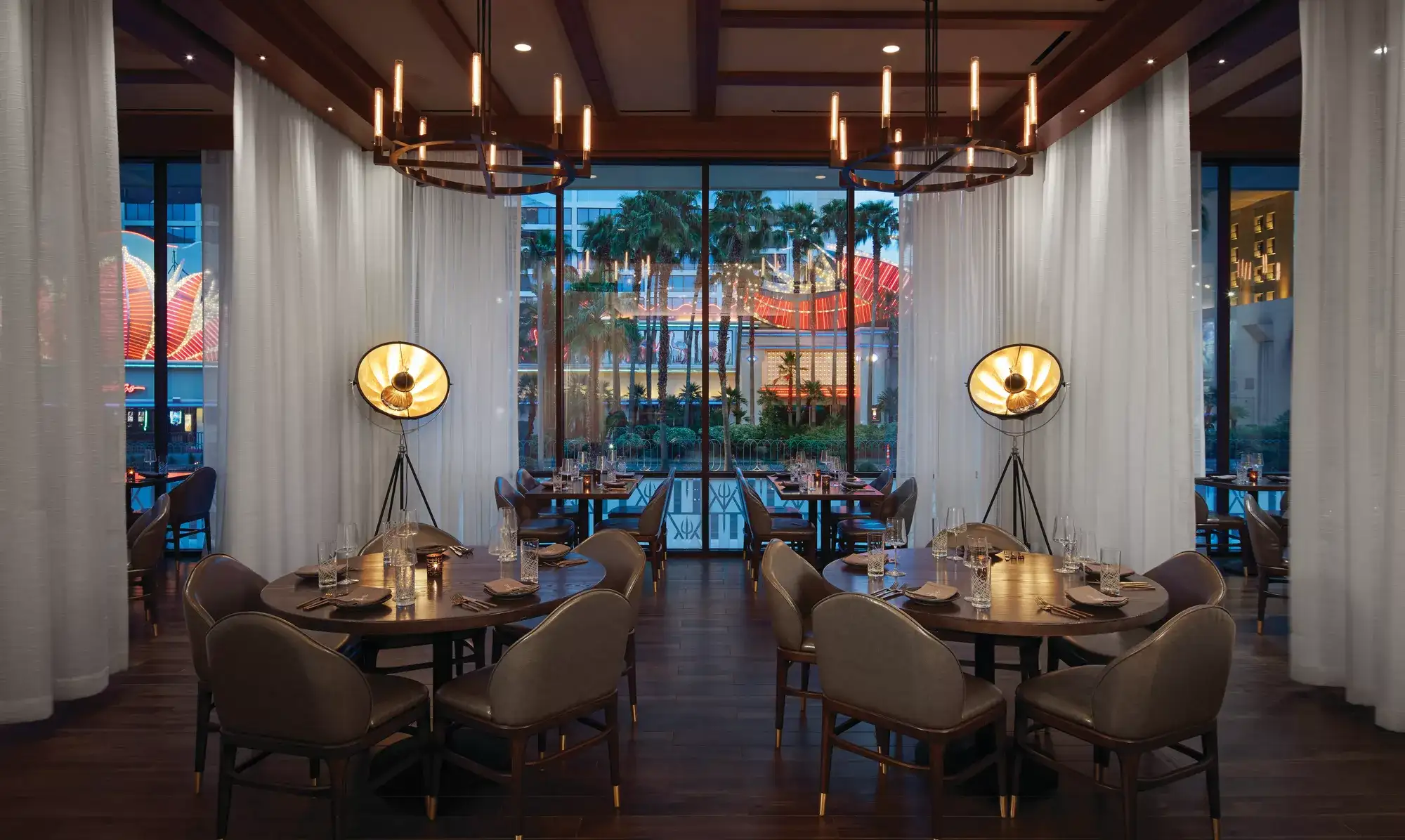 Upscale restaurant dining room with round tables and floor-to-ceiling windows overlooking a city street.