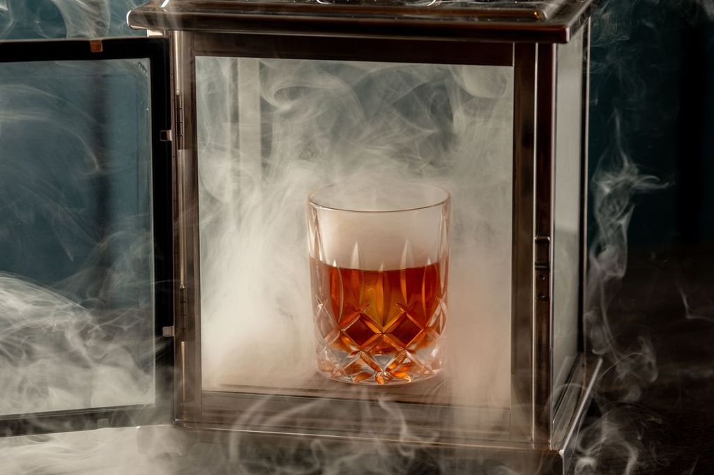 A glass of amber liqueur inside a metal lantern filled with smoke.