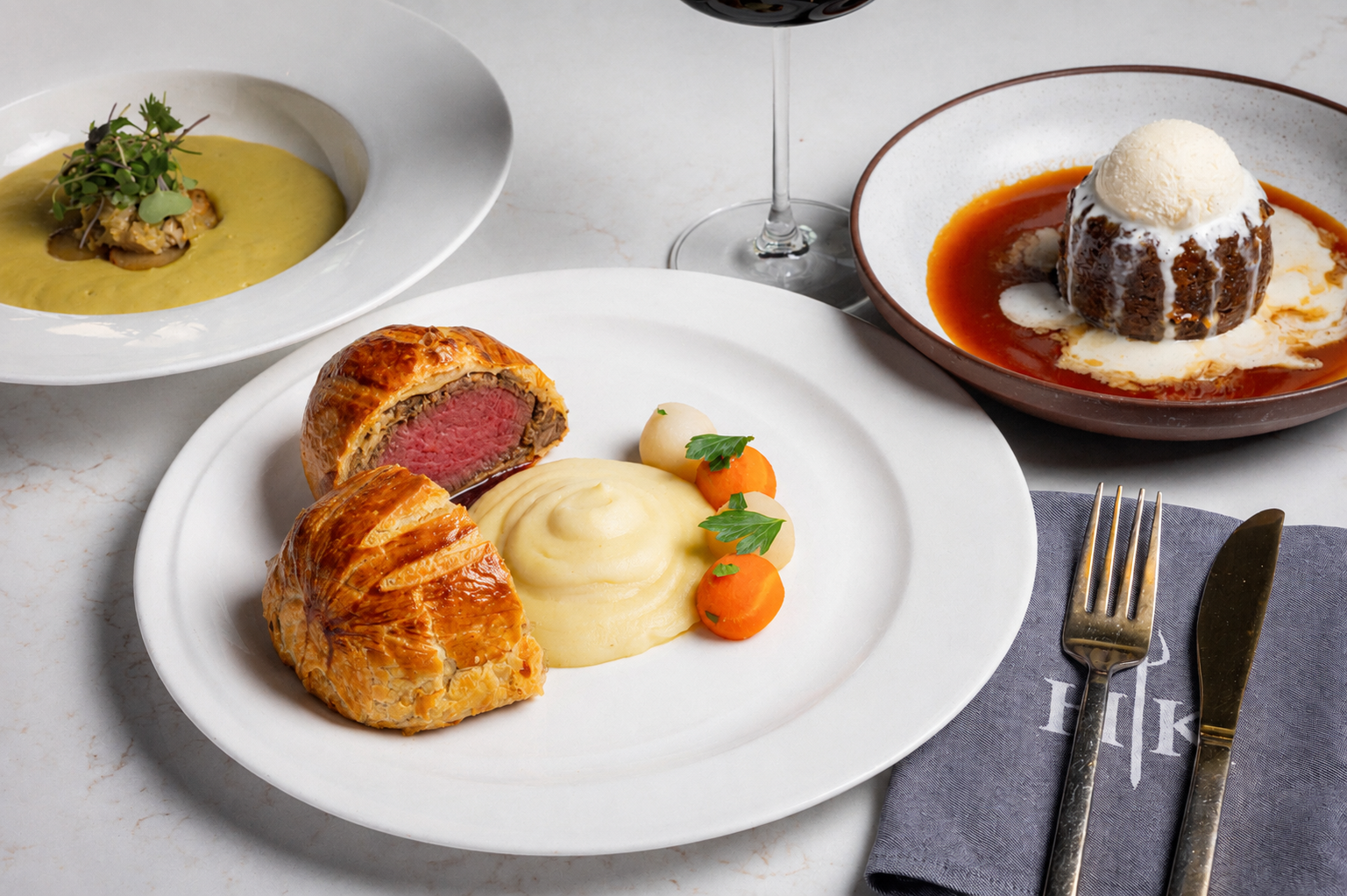 Beef Wellington with mashed potatoes and vegetables, flanked by soup and a pudding, with cutlery on a serviette.
