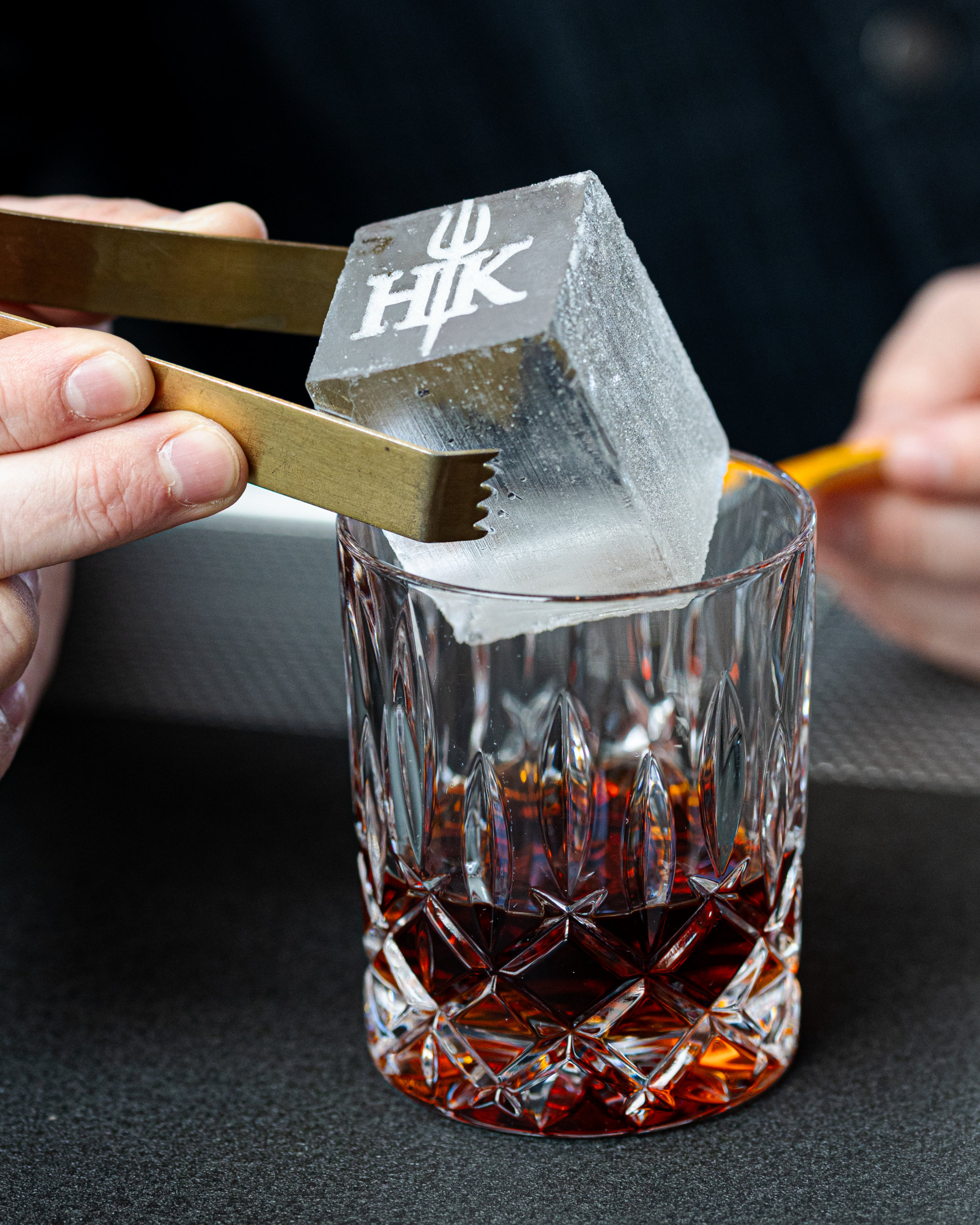Brass tongs lowering a large ice cube stamped with a logo into a crystal glass filled with amber liquid.