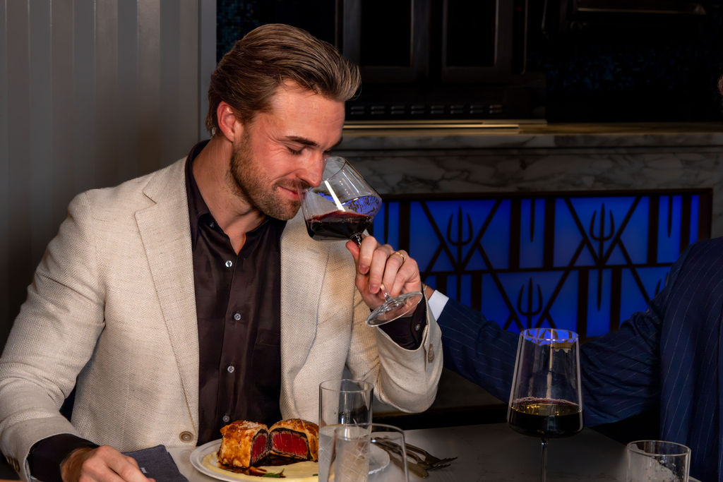 A man in a blazer smelling a glass of red wine at a table with a beef Wellington.