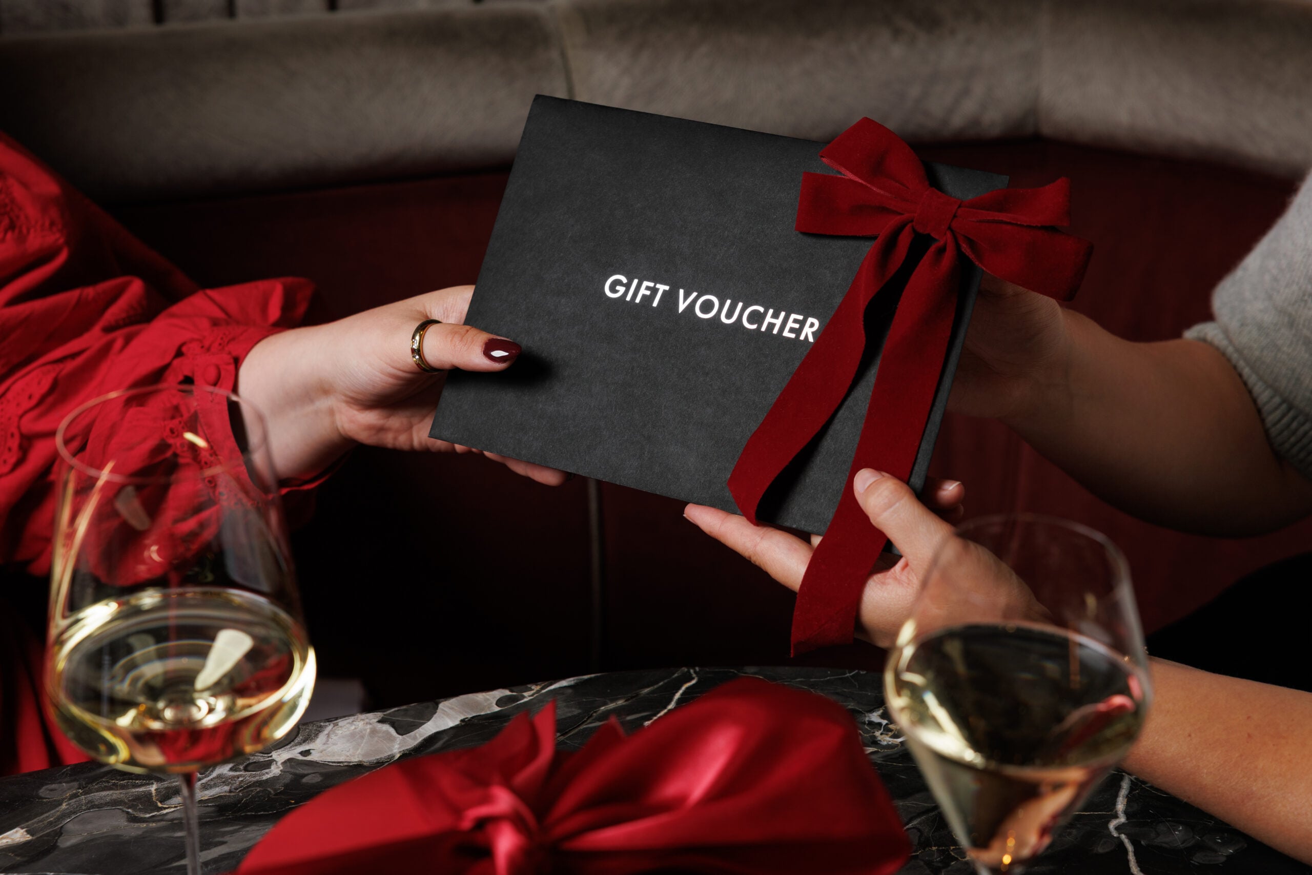Two people holding a black gift voucher with a red velvet bow.