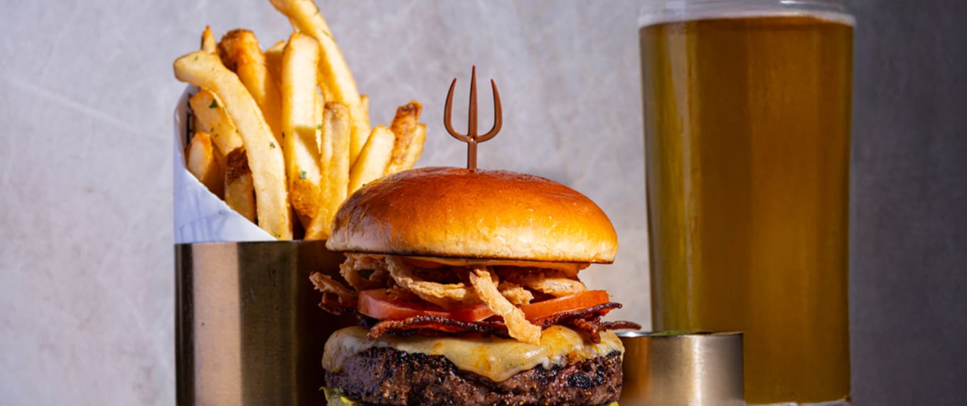 Cheeseburger with bacon, crispy onions, chips in a cup, and a pint of beer on a table.