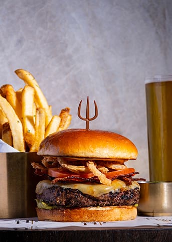 Cheeseburger with bacon, crispy onions, and tomato, served with chips and a glass of beer in the background.