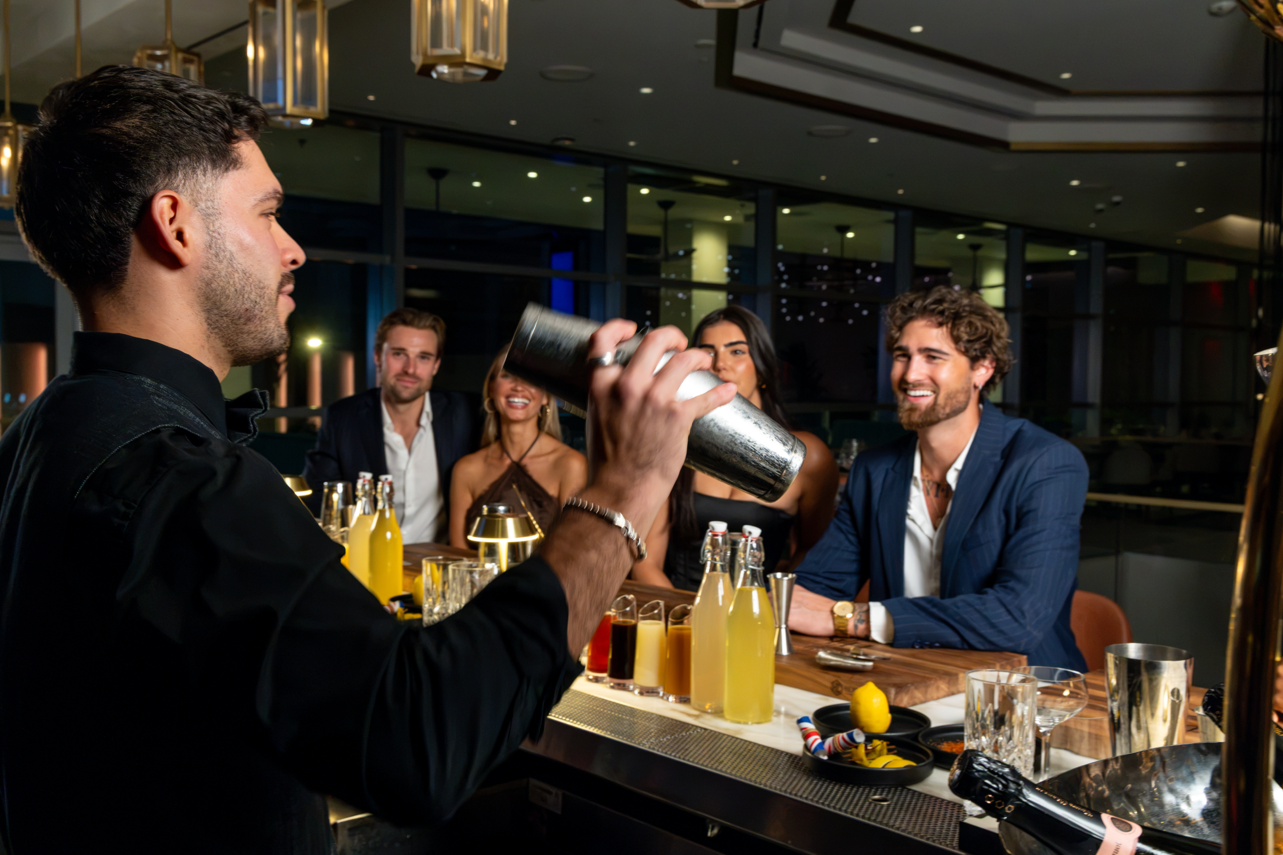 A barman shaking a cocktail at a bar with three customers watching and smiling.