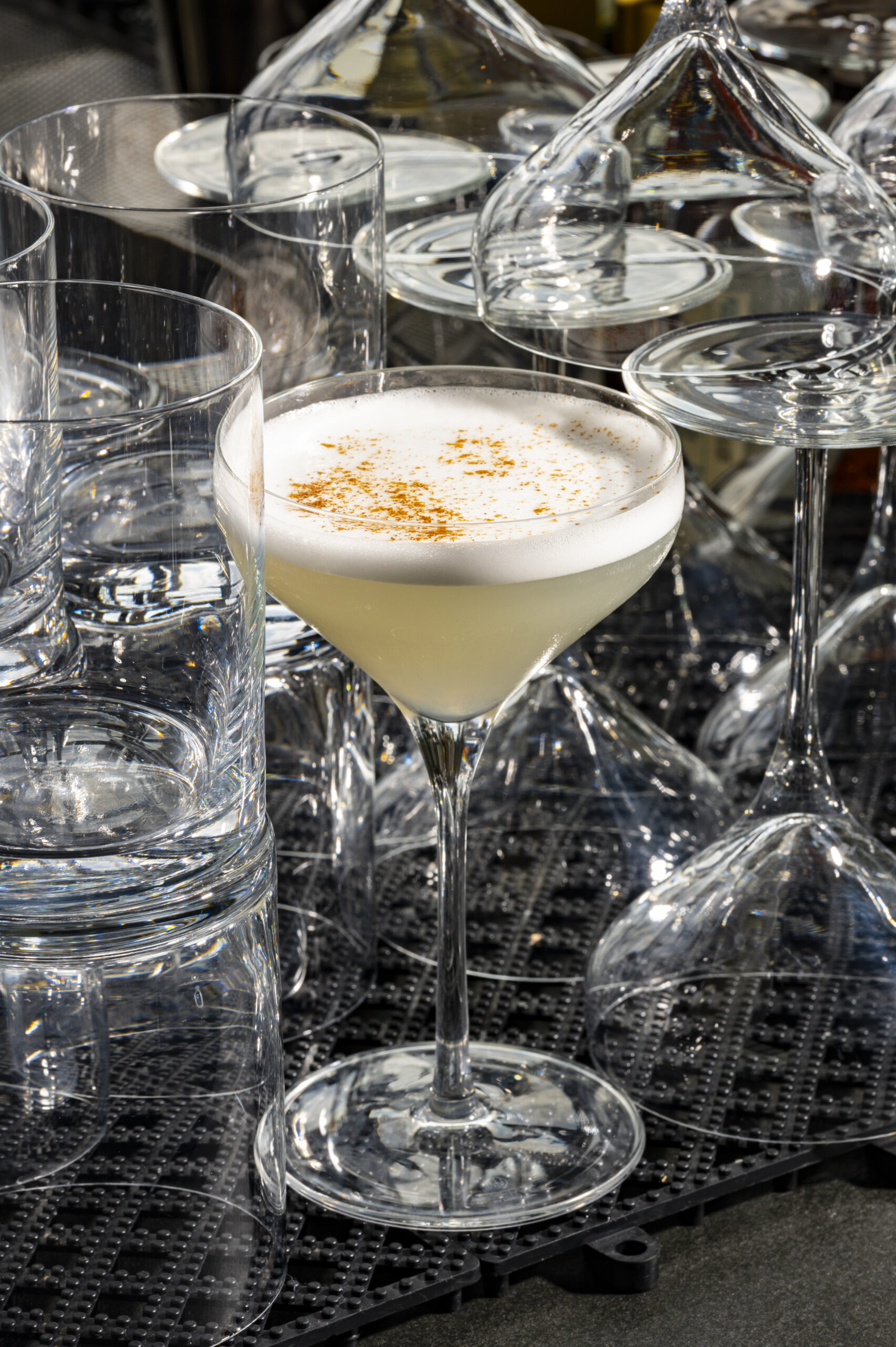 A martini glass filled with a pale cocktail topped with foam and sprinkled spice, surrounded by empty glassware.