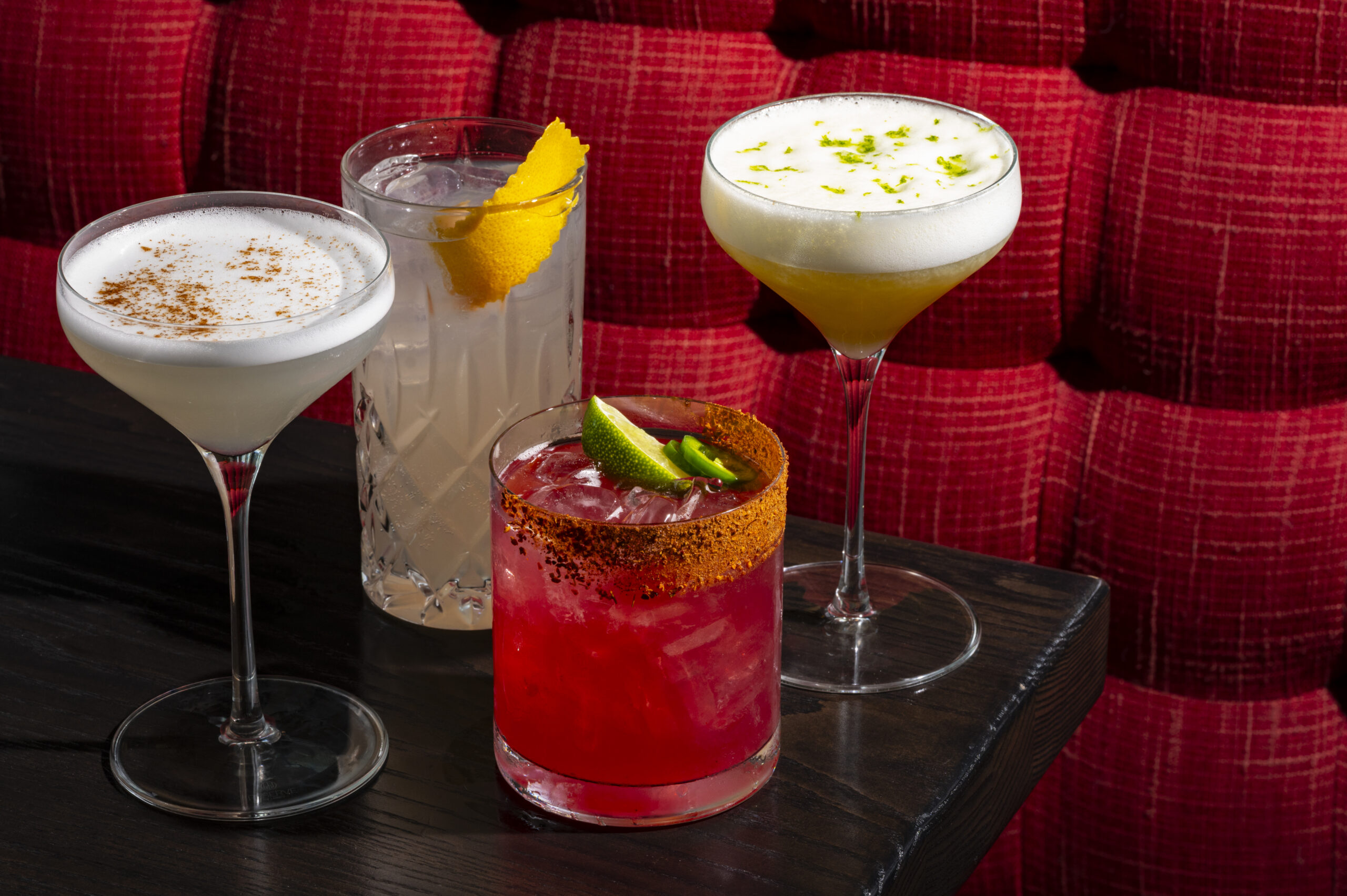 Four different cocktails on a dark wooden table against a red upholstered background.