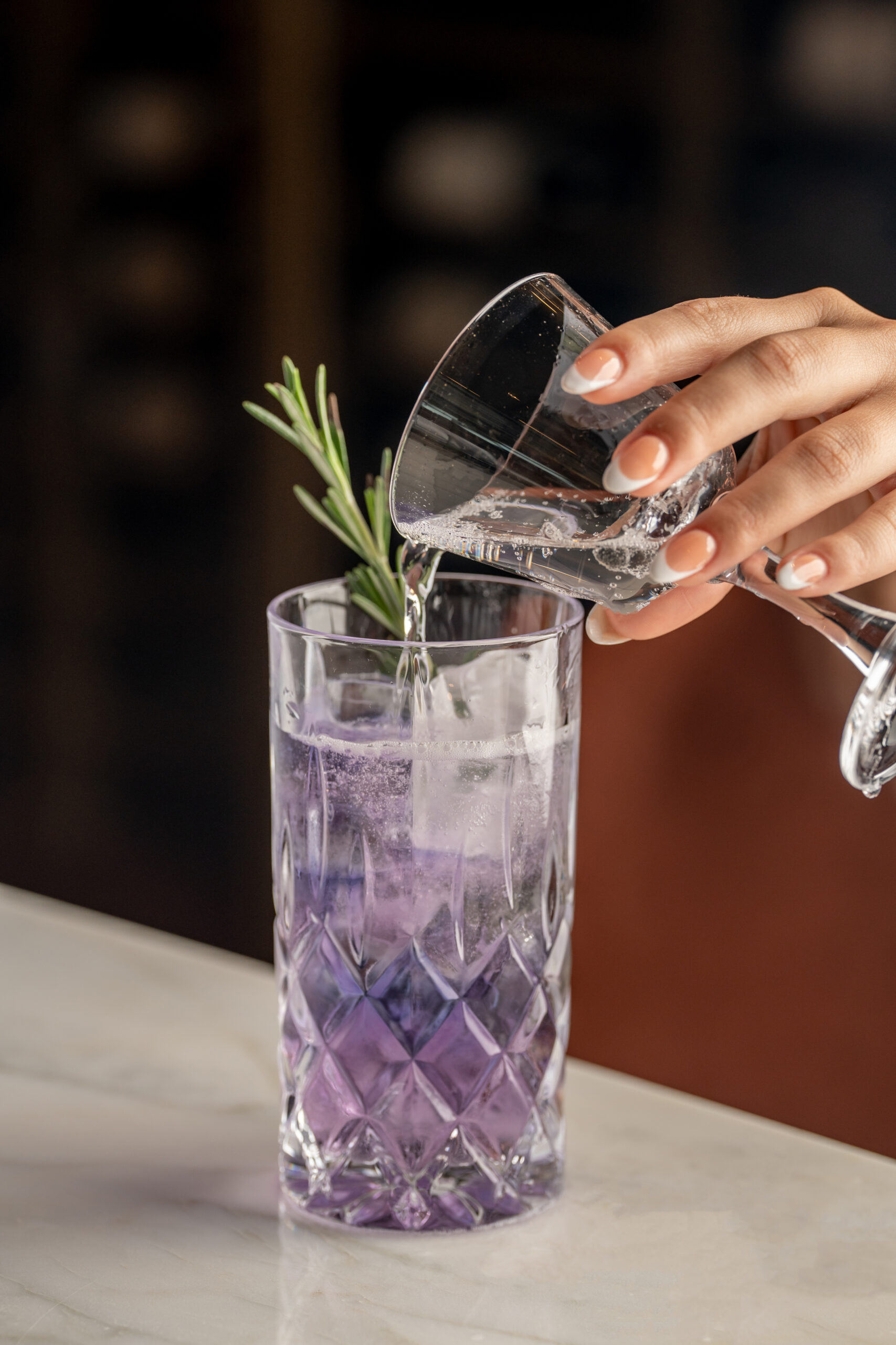 A hand pours liquid from a small glass into a tall, patterned glass filled with purple liquid and a rosemary sprig.