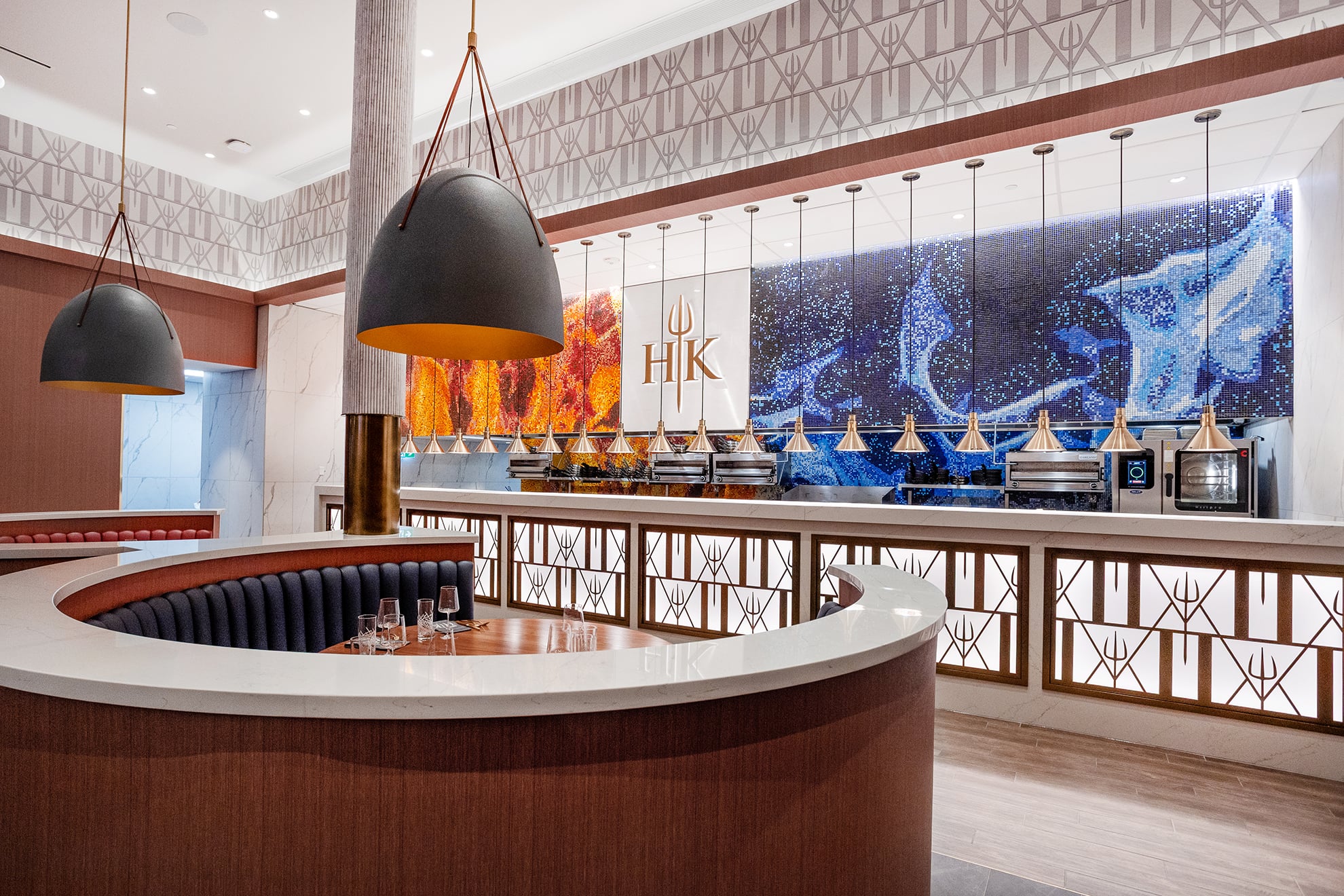 A semi-circular restaurant banquette facing an open kitchen with an illuminated “HK” logo and a blue tiled mosaic wall.