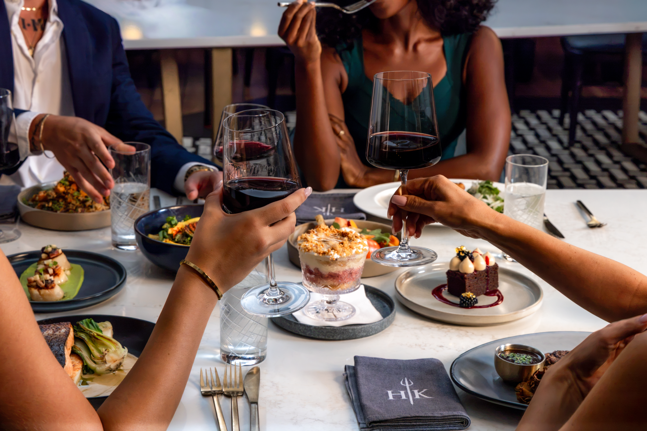 People raise glasses of red wine in a restaurant, with various dishes and puddings on the table.