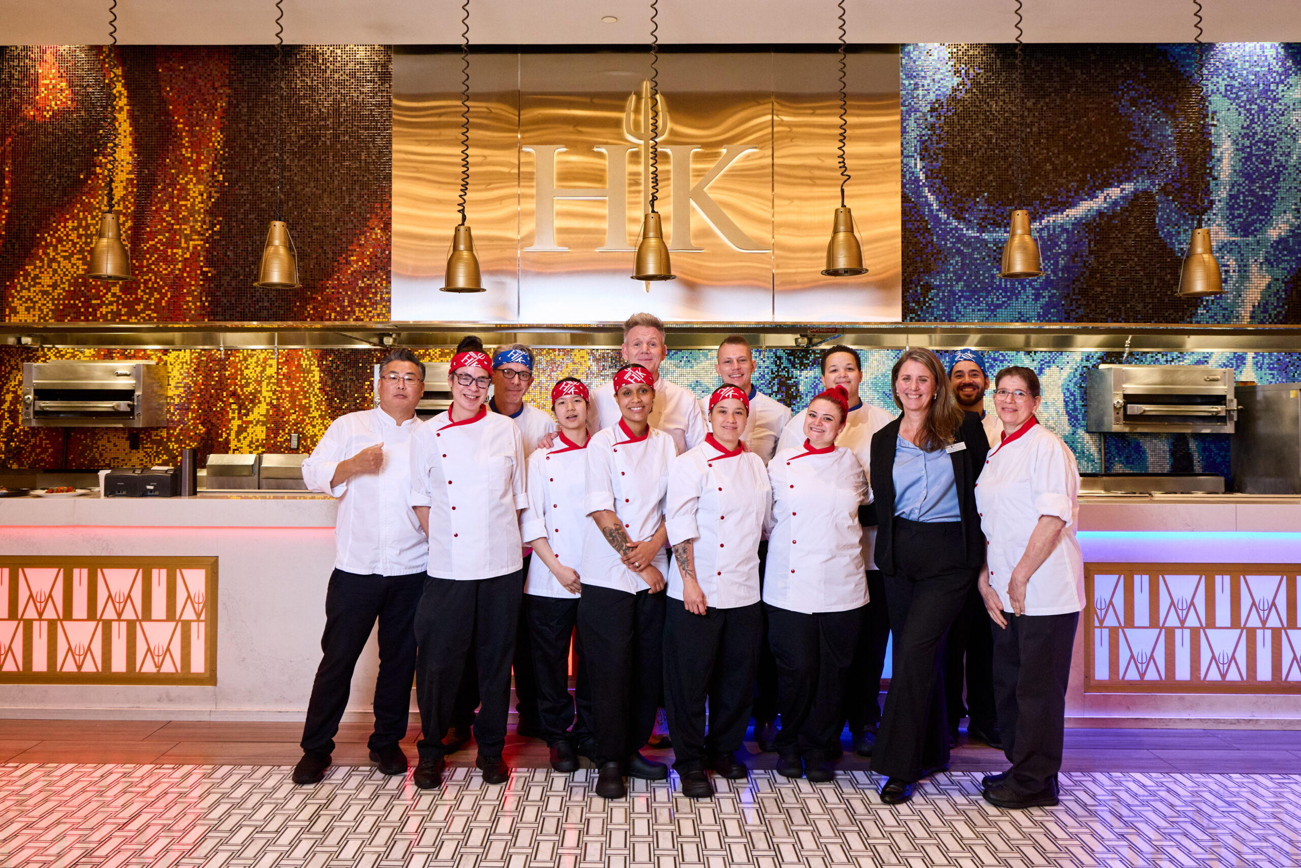 Gordon Ramsay posing with a group of chefs in Hell’s Kitchen uniforms in front of a branded kitchen counter.