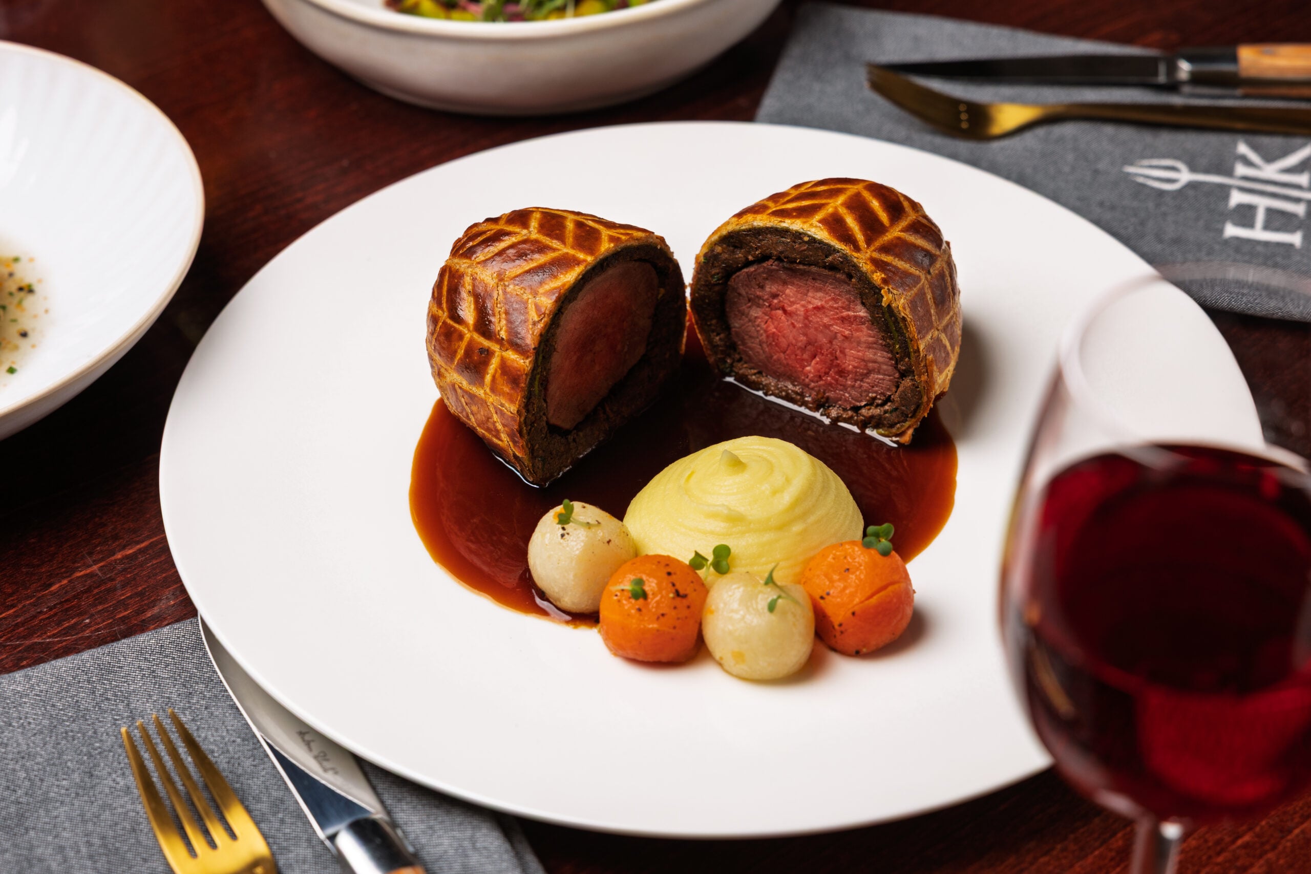 Beef Wellington sliced in half, with mashed potatoes, carrots, swedes, and red wine on a white plate.