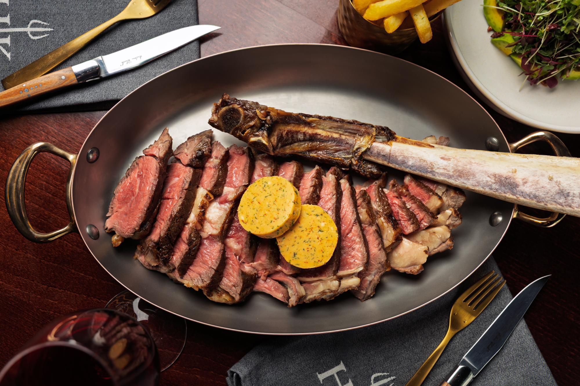 Sliced tomahawk steak topped with two herb butter rounds in a serving dish.