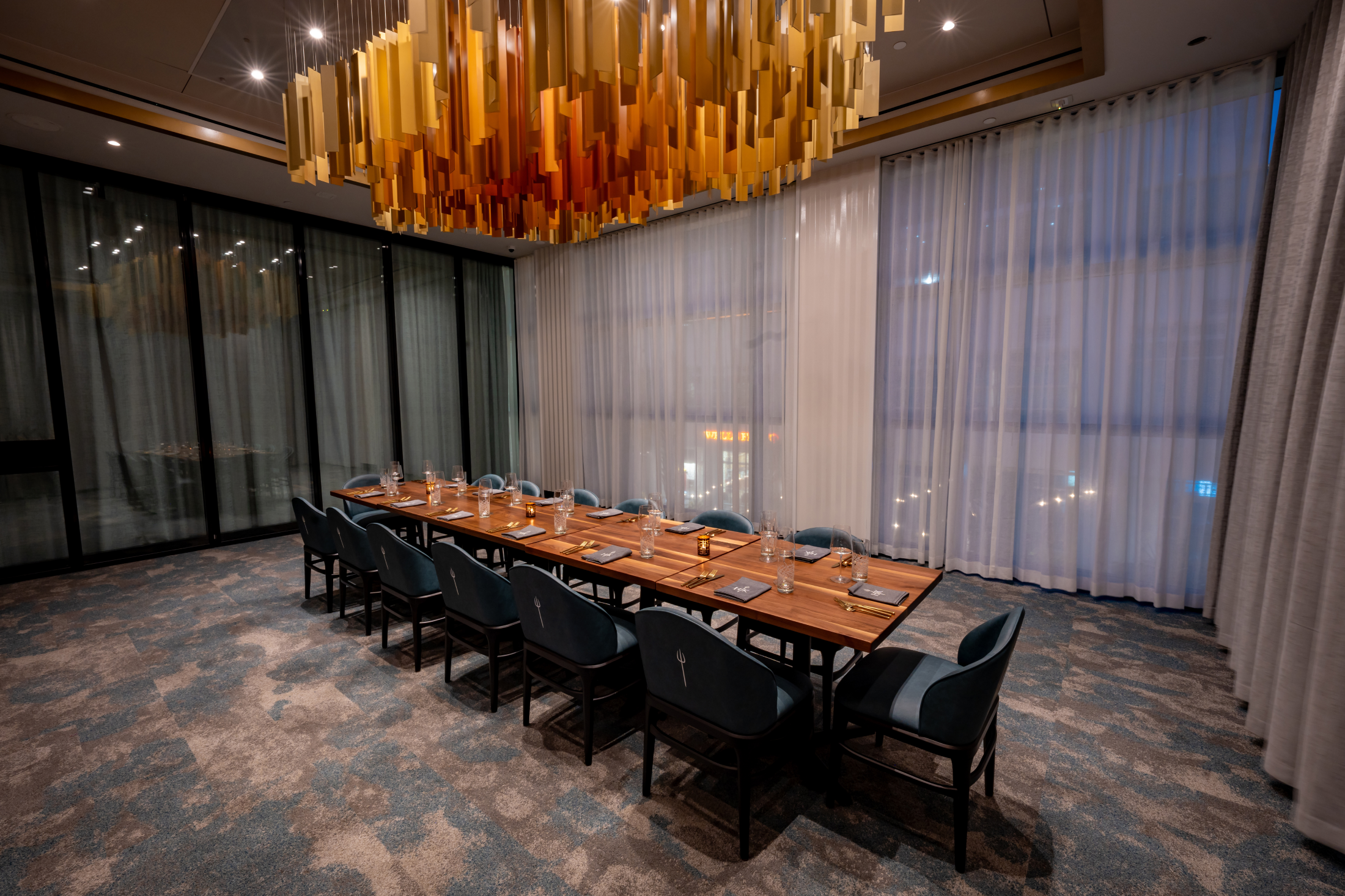 Long rectangular dining table with place settings, surrounded by blue chairs under a gold hanging light fitting.