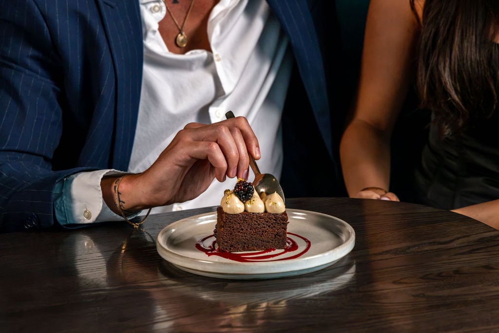 A person in a suit eats a slice of chocolate cake from Hell’s Kitchen, topped with cream and a blackberry.