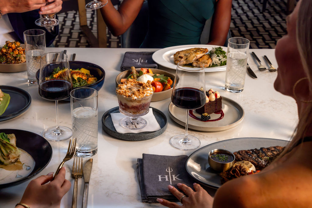 People at a Hell’s Kitchen restaurant table with various main courses, desserts, and glasses of red wine.
