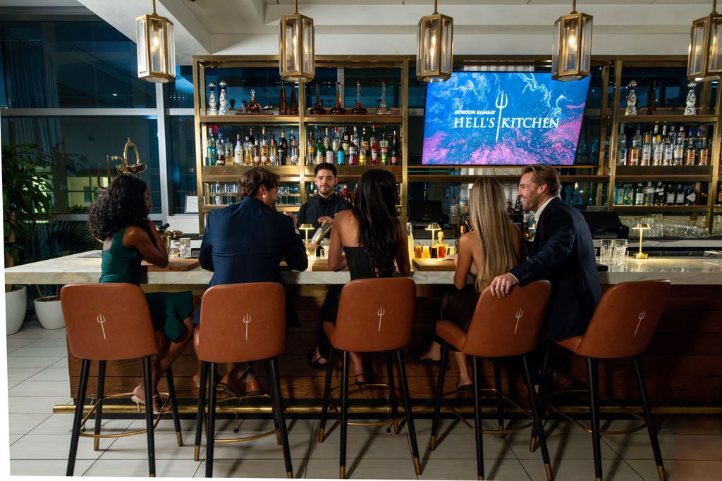 People sitting at a bar with a Hell’s Kitchen logo displayed on a television screen.
