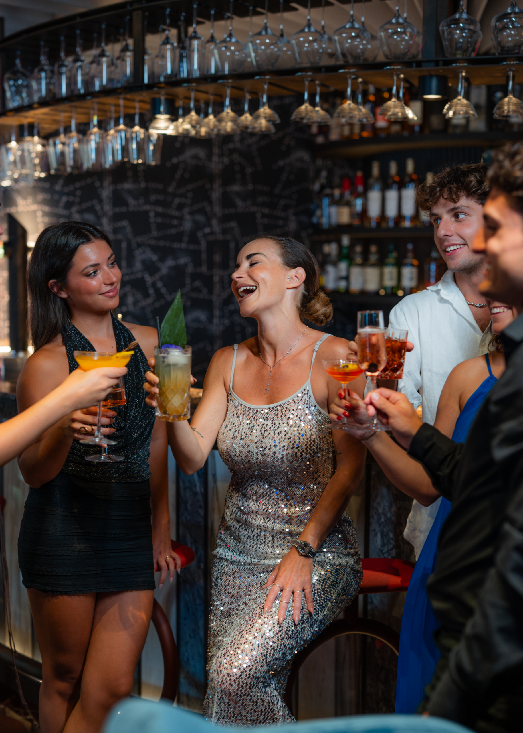Group of people dressed up, laughing and toasting drinks at a lively bar or party.