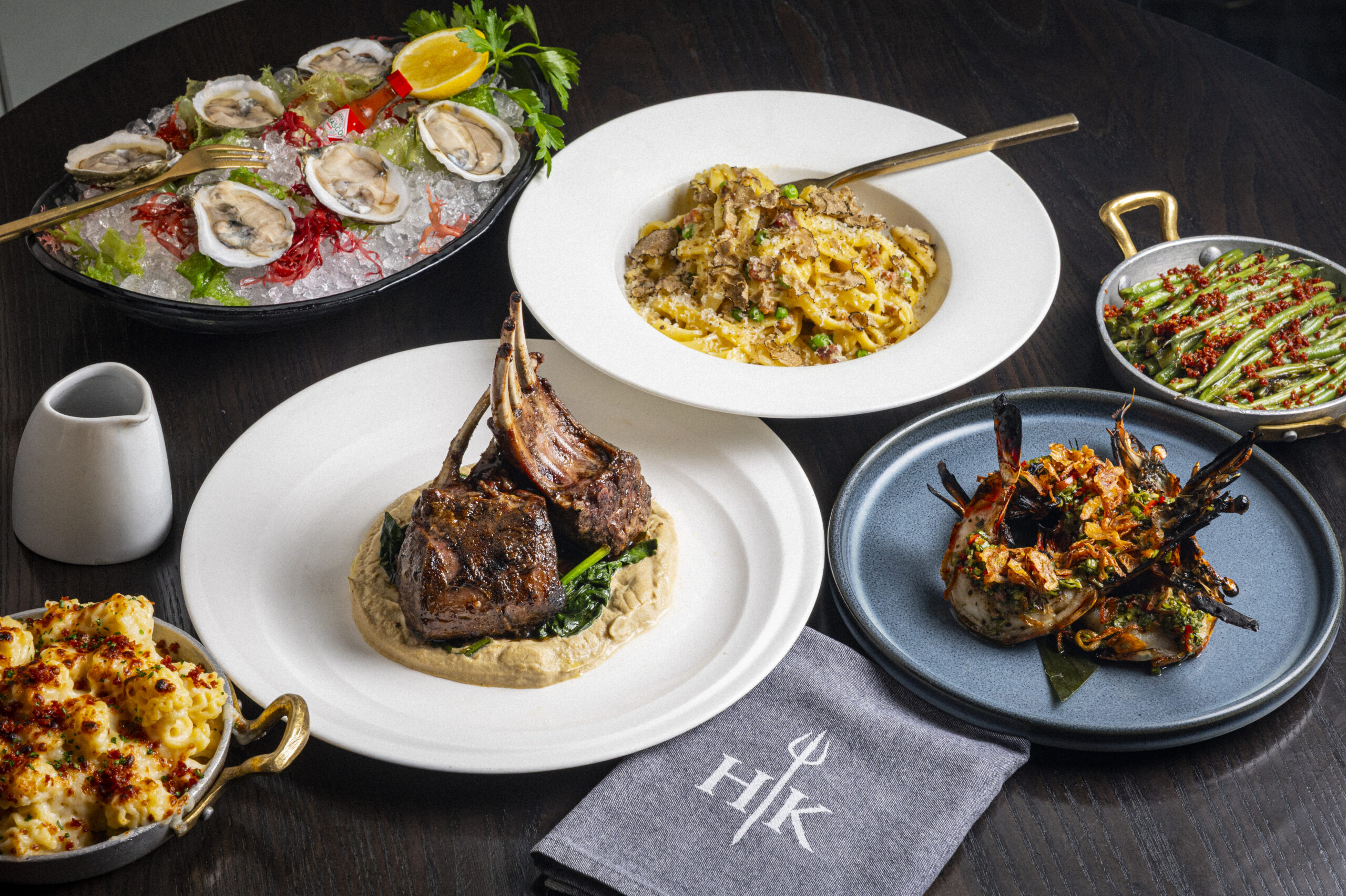 A spread of various gourmet dishes, including lamb chops, pasta, oysters, prawns, and sides on a dark table.