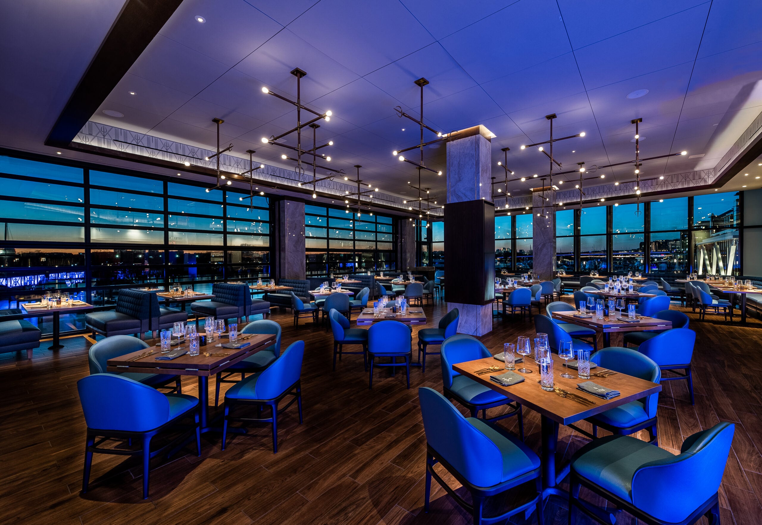 Modern Hell’s Hideaway Private Dining interior with blue chairs and wooden tables overlooking a waterfront at dusk.