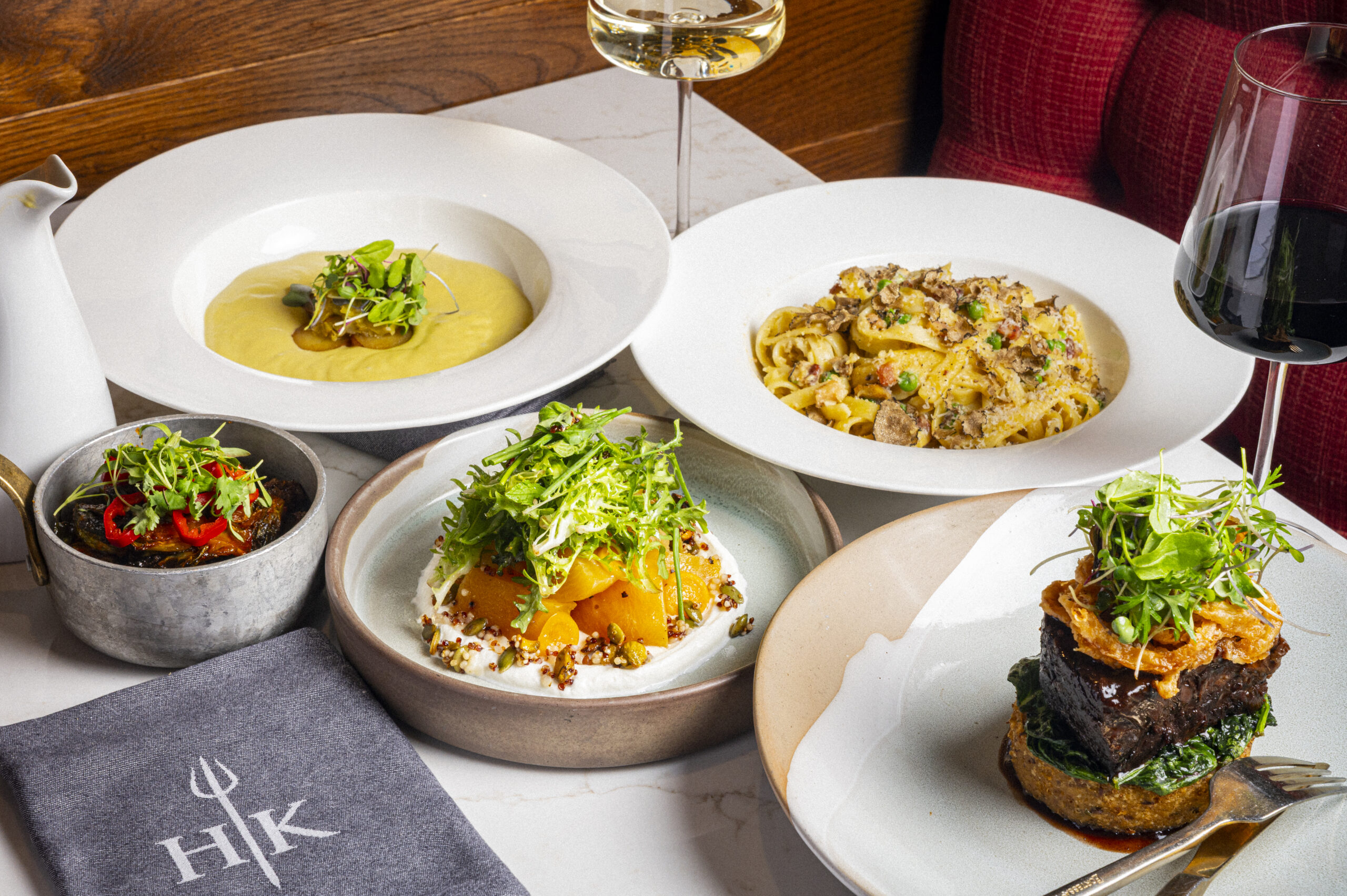 A table spread featuring bowls of pasta, soup, roasted squash, a beef main course, and glasses of wine.