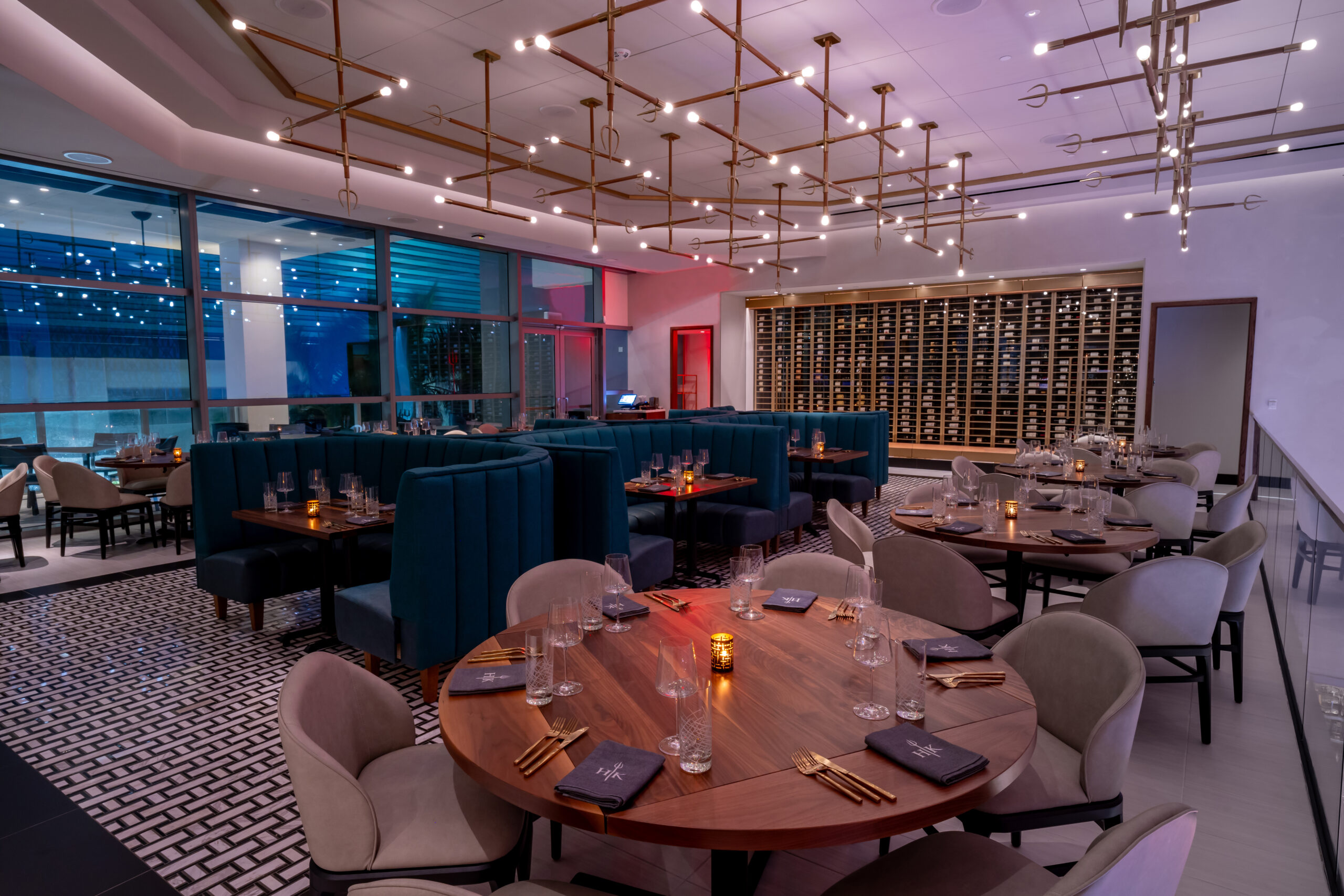 A restaurant dining room with blue curved banquettes, round wooden tables, and a large wine cellar wall.