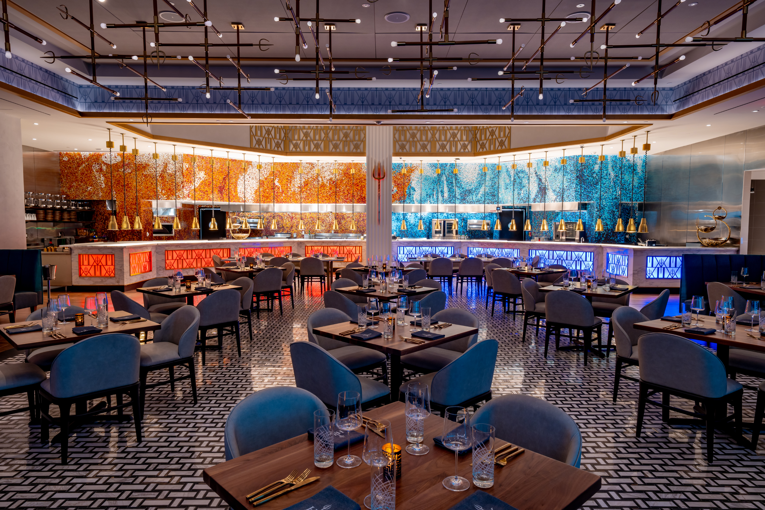 A modern restaurant dining room featuring tables with place settings, gray chairs, and a mosaic-tiled serving counter.