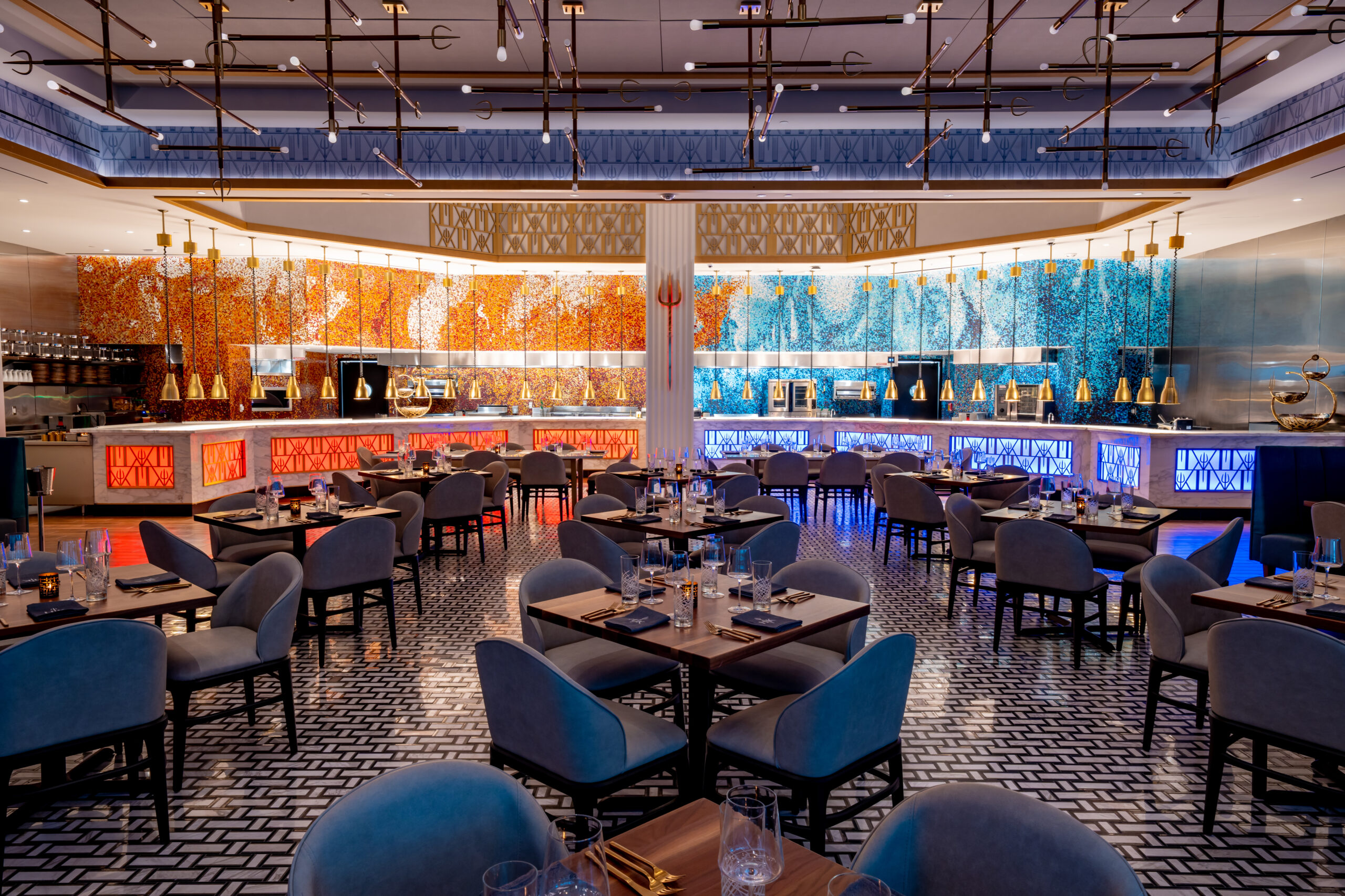 Dining room with rows of square tables, blue upholstered chairs, and a long counter with orange and blue mosaic tiles.