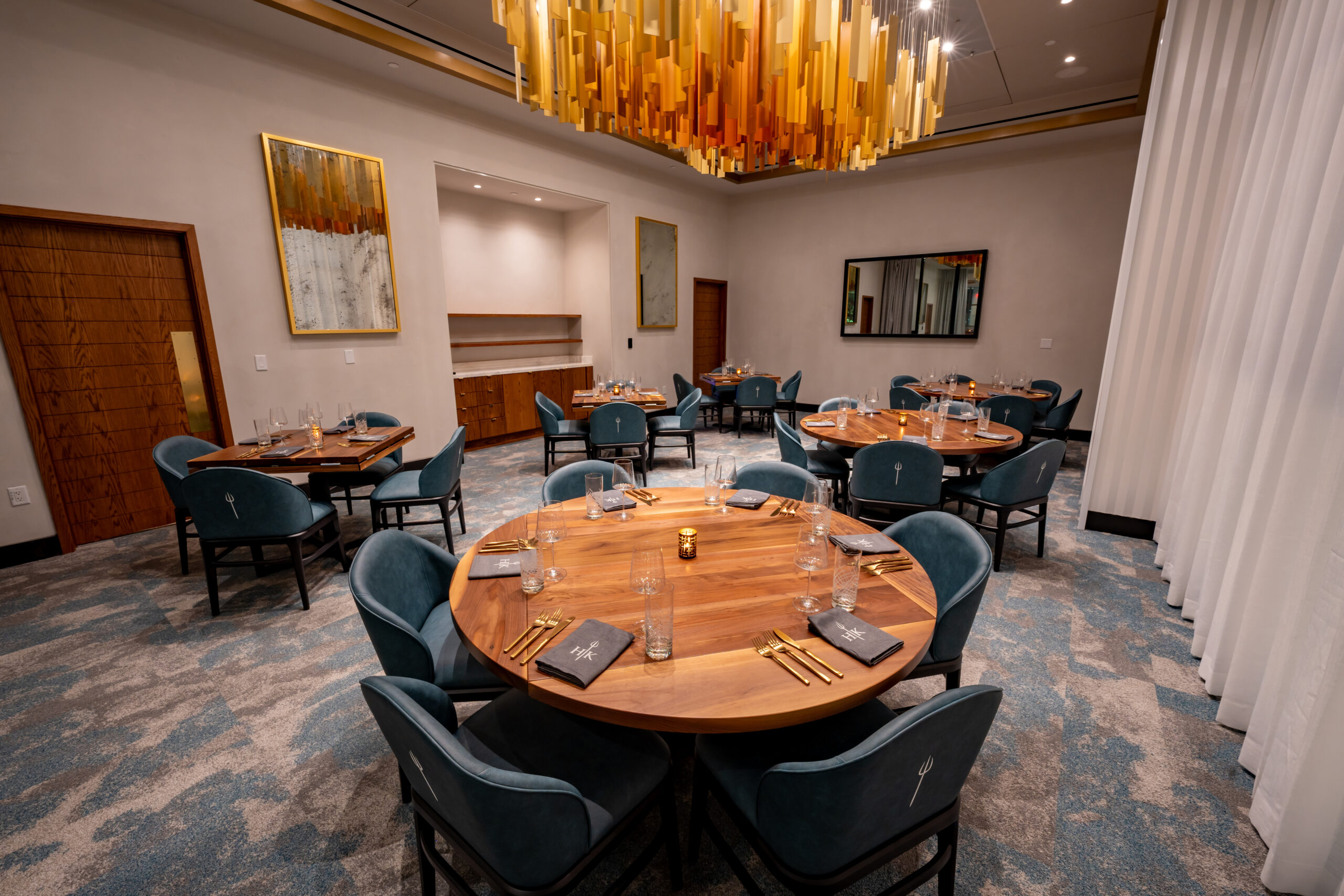 A dining room with round and square wooden tables set with blue chairs and a large amber-coloured modern chandelier.