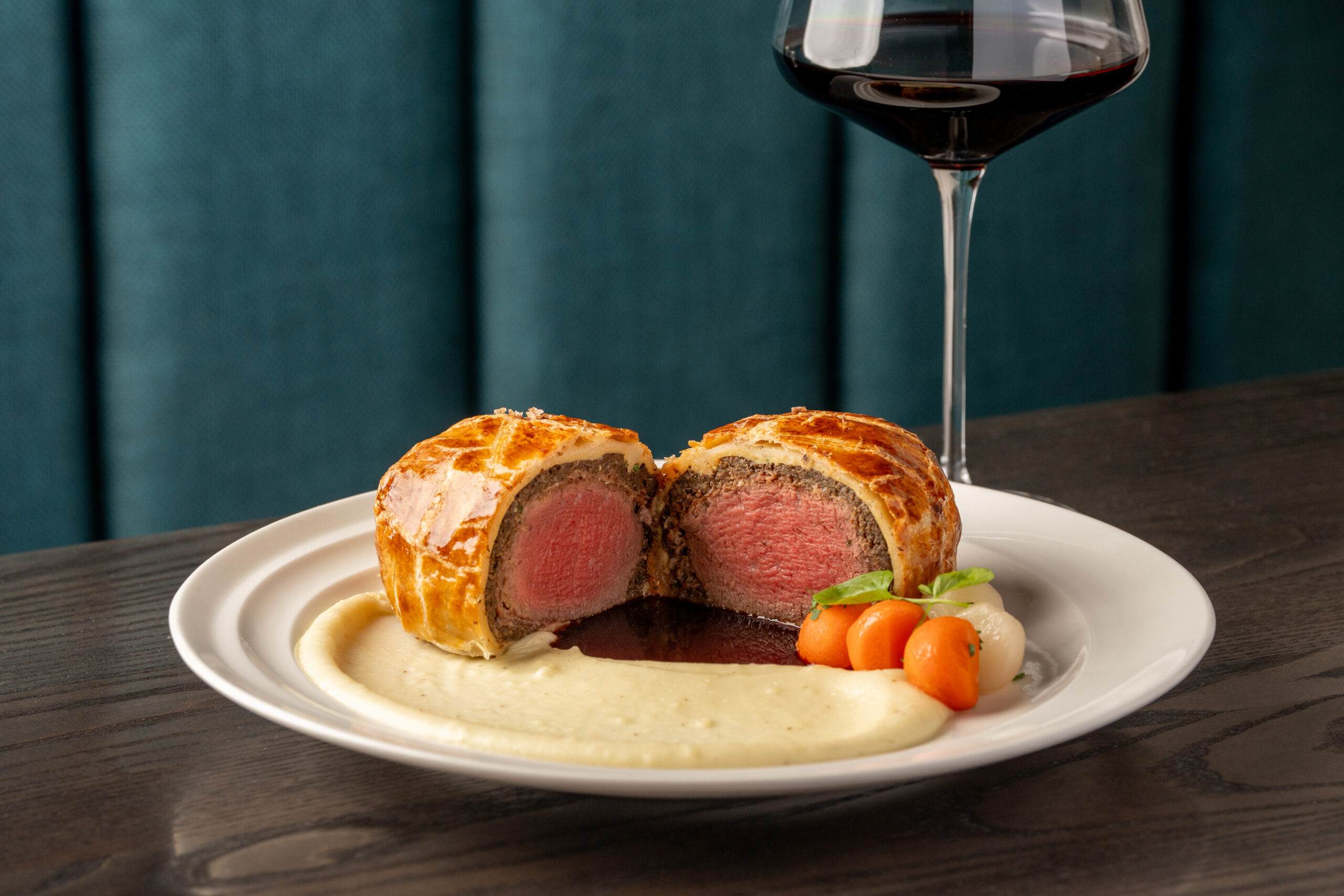 Beef Wellington sliced in half on a plate with puréed potatoes, glazed carrots, and a glass of red wine.