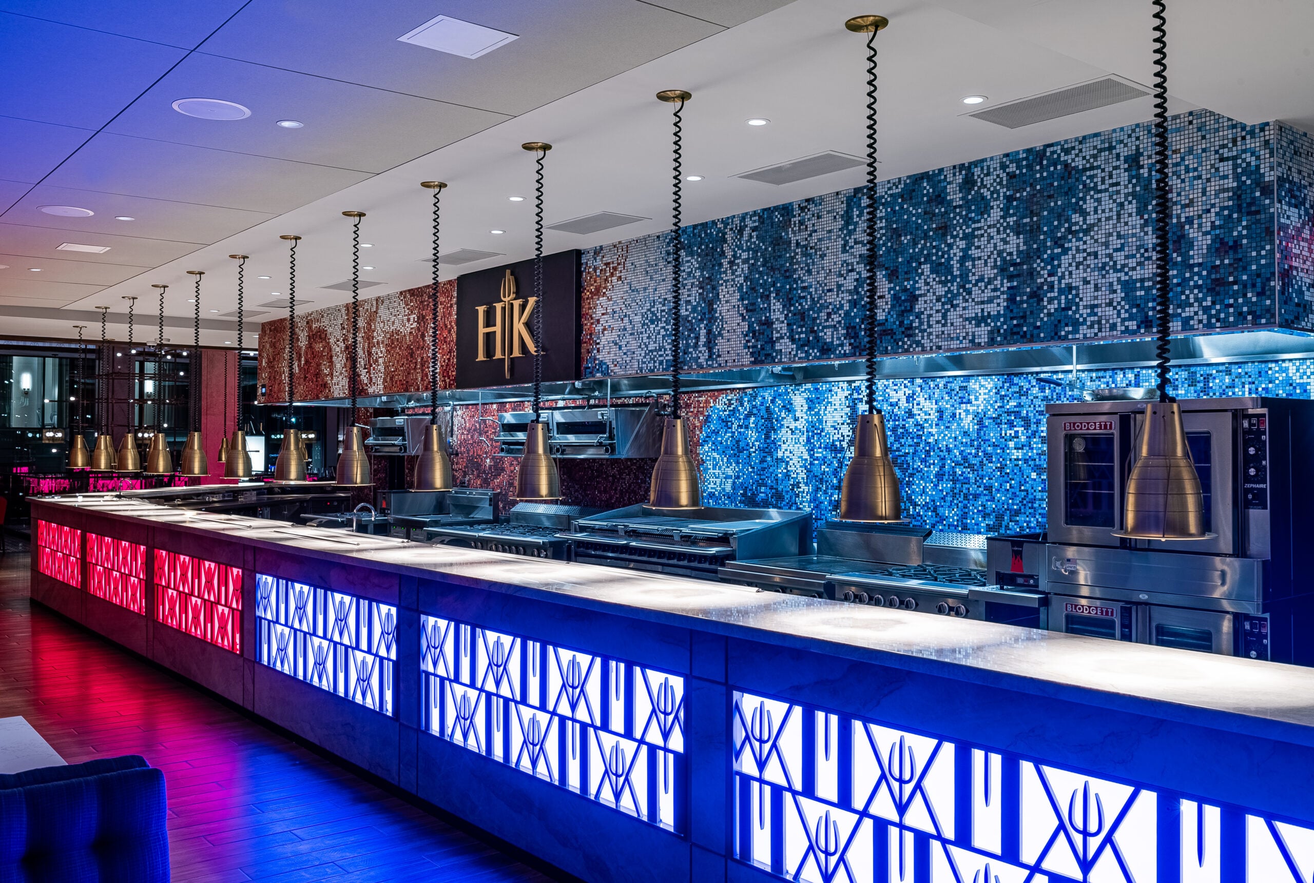 A modern Hell’s Hideaway Private Dining kitchen with a long counter under blue and red light and hanging heat lamps.