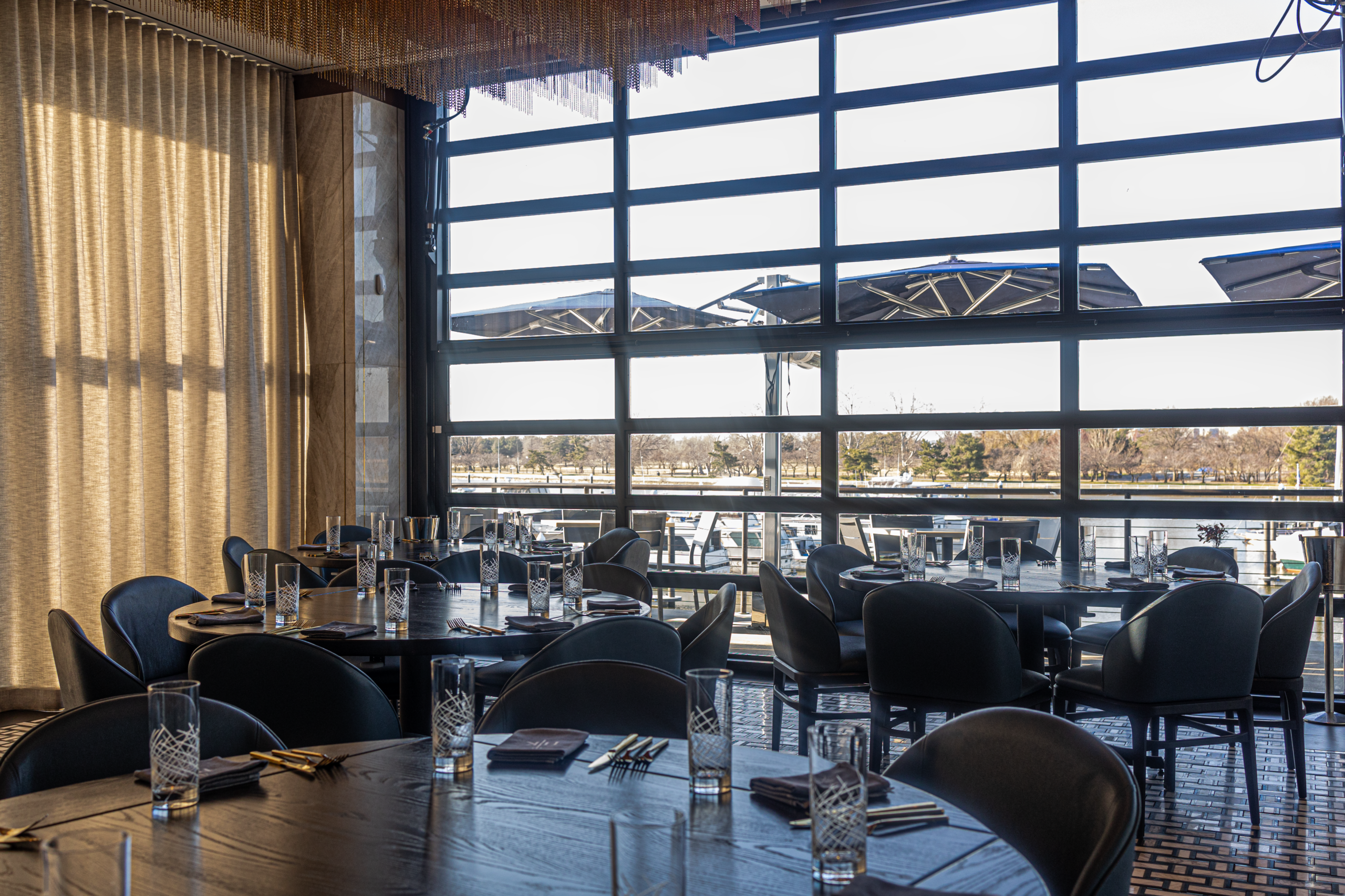 A restaurant dining room with dark tables and chairs, featuring large floor-to-ceiling windows overlooking a harbour.